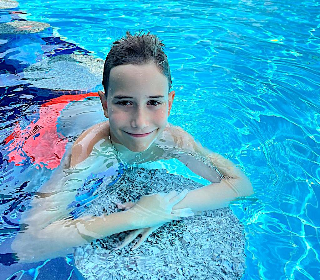 A smiling boy resting on the edge of a bright blue swimming pool